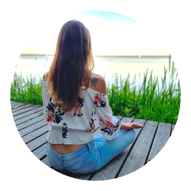 Jessica Wells sitzt im Schneidersitz auf einem Holzsteg am Wasser und blickt aufs Ufer